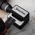 A SharpTool laser-etched steel square with a black and silver body, shown with a drill bit guide and part of a cordless drill on a worn, scratched white workbench surface.